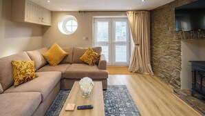 A living room with a beige sectional sofa, a round window, a stone wall, and a fireplace. - Bay Tree Cottage Rhondda Valley by STAE-Homes (Rhondda Cynon Taff)