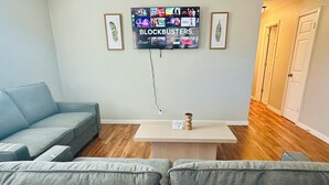 Una sala de estar con un sofá gris, una mesa de centro y un televisor montado en la pared mostrando 'BLOCKBUSTERS'.