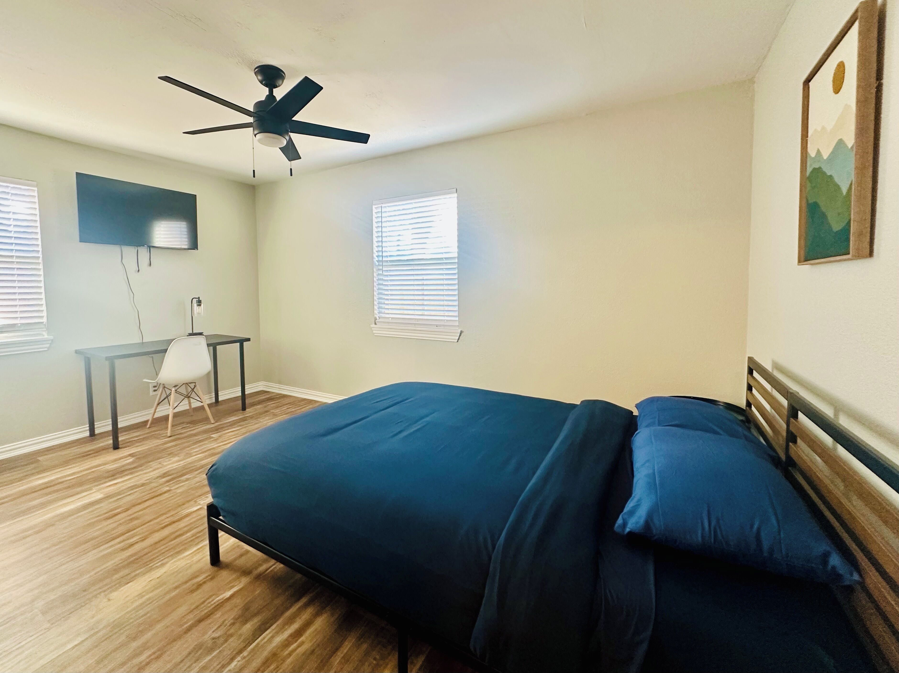 Un dormitorio con cama, escritorio, silla, ventilador de techo y dos ventanas con persianas.