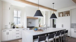 Eine moderne Küche mit weißen Schränken, einer großen Kücheninsel mit Barhocker, Pendelleuchten und Blick ins Freie.
