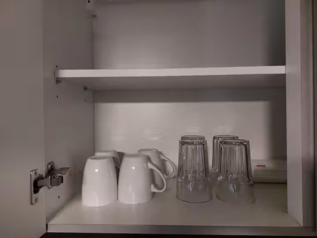 A white cabinet with two shelves; the lower shelf holds three white mugs and two clear glass mugs.
