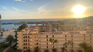 Un paisaje urbano costero con un edificio destacado que tiene varios balcones y un puerto deportivo al fondo.