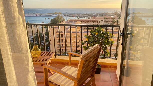 Un balcón con una silla y una mesa de madera, una planta en maceta y vistas al mar y edificios.