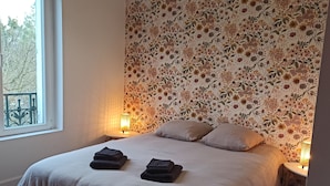 Ett sovrum med en blommig tapet, en säng med två kuddar, två sänglampor och ett fönster med utsikt över träd.