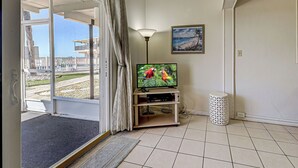 A room with a sliding glass door leading to a balcony, a television on a wooden stand, a floor lamp, and a painting on the wall.