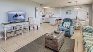 Una sala de estar con televisor, sofá, sillón, mesa de centro y una cocina al fondo.