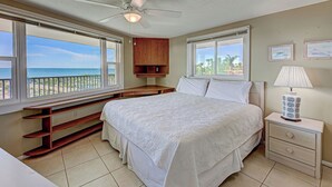 Un dormitorio con cama, mesita de noche, lámpara, ventilador de techo, ventana con vista a la playa y una estantería de madera.