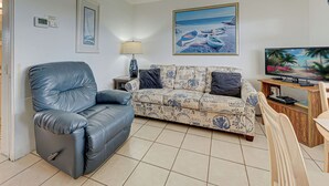 Un salón con un sofá de cuero azul, un sofá estampado, una mesita de madera, un televisor y un cuadro de barcos en la playa.