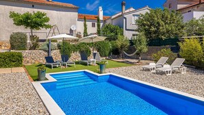 A swimming pool with lounge chairs, a stone wall, and a garden with trees and shrubs.