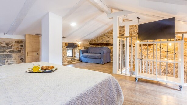 A cozy loft bedroom with a stone wall, a bed with a white blanket, a blue sofa, and a TV on a white stand.