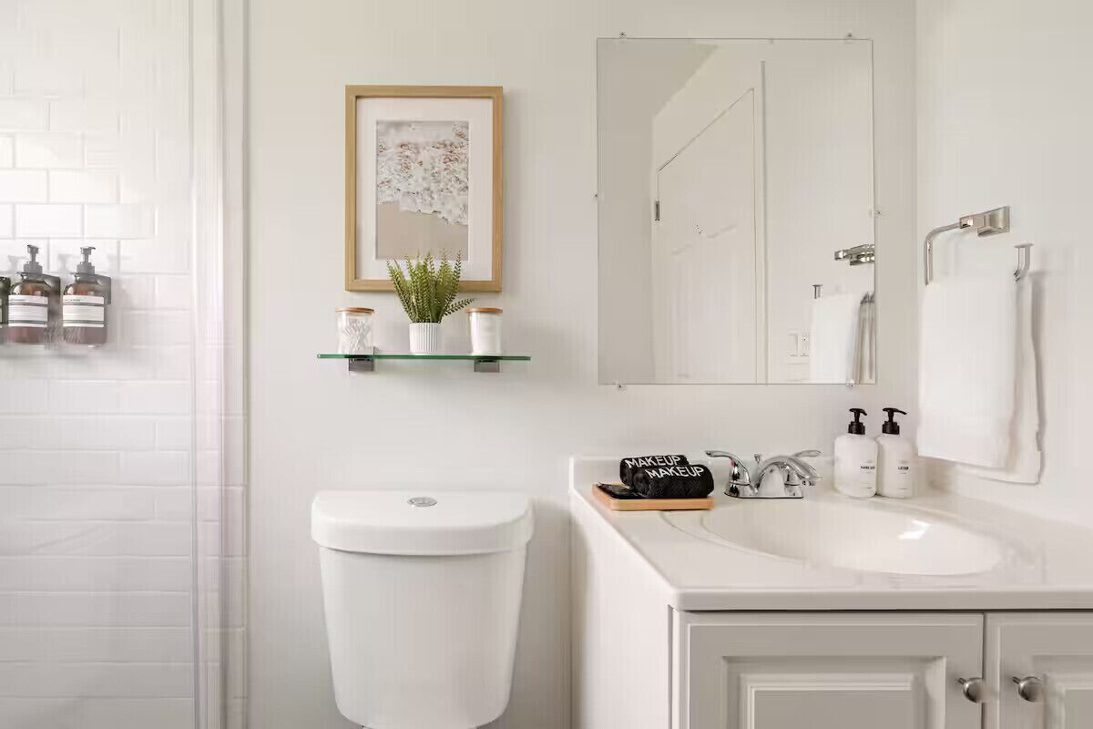 A modern bathroom with a toilet, sink, and mirror. There are shelves with toiletries and a framed picture on the wall.