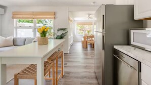 Una cucina moderna con un frigorifero in acciaio inox, una zona pranzo con tavolo e sedie bianche, e un'ampia finestra vista su del verde.