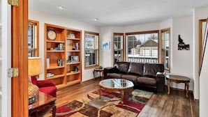 A living room with a brown leather sofa, a glass coffee table, a red armchair, wooden bookshelves, and a large window with blinds. - Moose Caboose Hot Tub + Schweitzer Views (Bonner County)