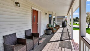 A covered porch with wicker chairs, a red door, and a view of a residential street. - Moose Caboose Hot Tub + Schweitzer Views (Bonner County)