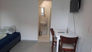 A compact room with a bed, a dining table with chairs, a TV mounted on the wall, and a bathroom with a sink and mirror. - Flat Bangoua (Abstatt)
