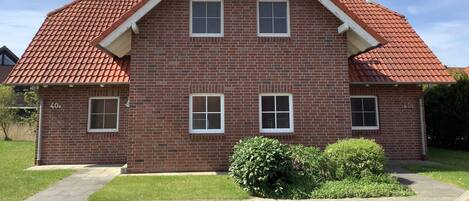 A two-story brick house with a red-tiled roof, two windows on the front, and a small front yard with a bush.