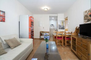 Un salon avec un canapé, une table basse ornée d’un vase, un coin repas avec des chaises, un téléviseur et une cuisine équipée d’un réfrigérateur.