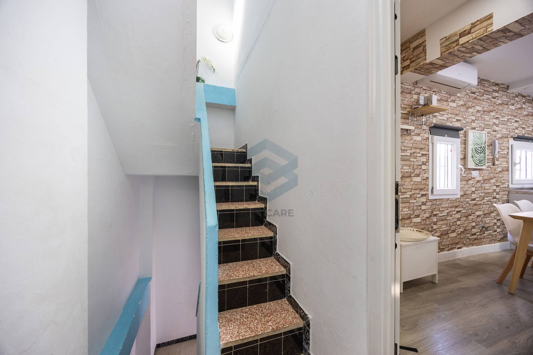 Una escalera con barandilla azul que lleva a una habitación con pared de piedra y una ventana.