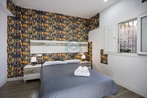 A hotel room with a bed, two bedside tables with lamps, a window with decorative screen, and a wall with a pattern of letters and symbols.
