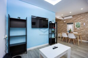 A modern living room with a TV, shelving units, a dining area with chairs, and a white table.