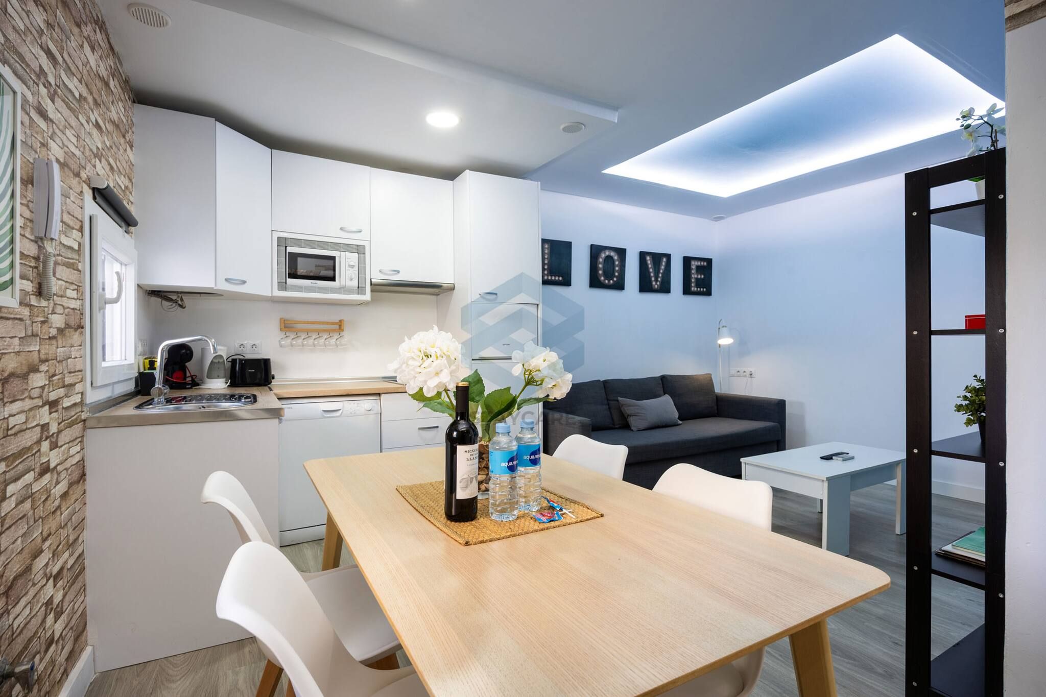 A modern kitchen with white cabinets, a microwave, and a coffee machine. There is a dining table with chairs and a vase of flowers. A blackboard with illuminated letters is on the wall.