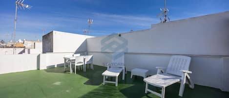 A rooftop terrace with white outdoor furniture, including chairs and a table, under a clear blue sky.