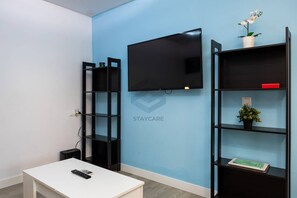 A room with a white table, a black TV mounted on the wall, and two black shelving units with various items on them.