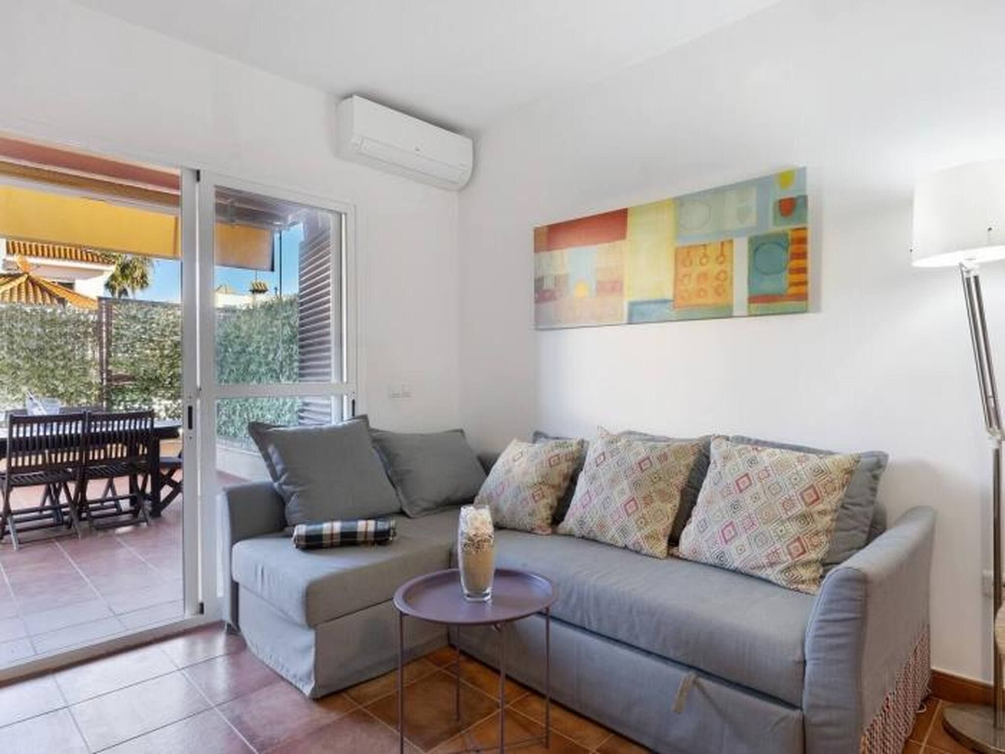 Una sala de estar con un sofá gris, cojines estampados, una pequeña mesa redonda y un cuadro abstracto colorido en la pared.