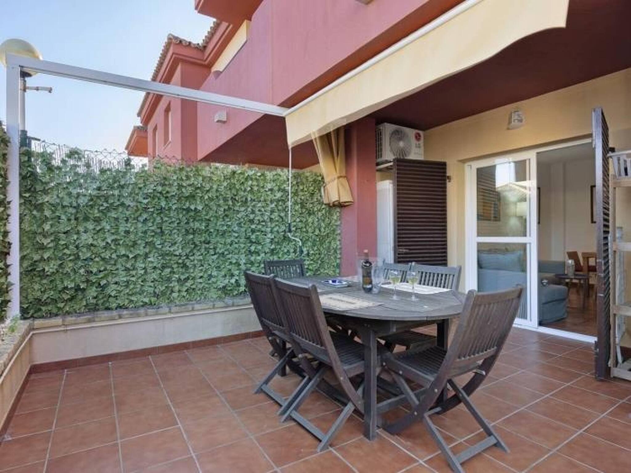 An outdoor patio with a table and chairs, a green hedge, and a building with a red facade.