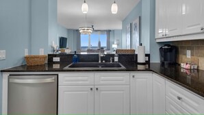A modern kitchen with white cabinets, a black countertop, and a stainless steel dishwasher. There are two pendant lights and a chandelier above the sink.