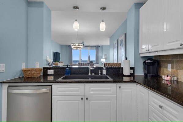 A modern kitchen with white cabinets, a black countertop, and a stainless steel dishwasher. There are two pendant lights and a chandelier above the sink. - Beach Colony West 11E / Gulf Front w/Beach Service & Onsite Management (Navarre Beach)