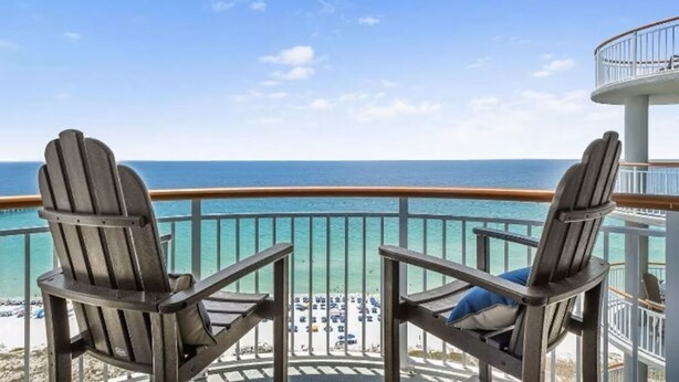 Un balcon avec deux fauteuils Adirondack donnant sur une plage aux eaux turquoise et limpides, avec un bâtiment blanc en arrière-plan.