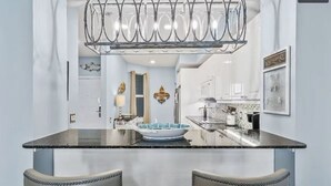 Une cuisine moderne dotée d’un îlot central, de deux chaises rembourrées, d’un luminaire décoratif en métal et d’une pancarte d’accueil.