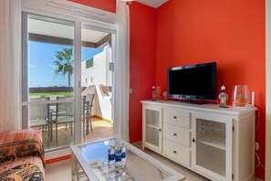 Ein Zimmer mit Fernseher auf einer weißen Kommode, einem Glastisch mit Wasserflaschen und Blick auf einen Balkon mit Gartenmöbeln.