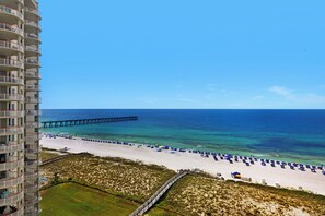 A beach with a pier extending into the ocean, a sandy beach with umbrellas, and a high-rise building with balconies.