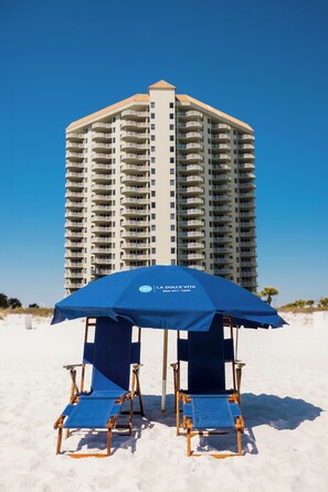 A beach umbrella with a logo, two blue lounge chairs, and a tall hotel building in the background.