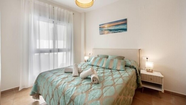 A neatly made bed with a patterned bedspread, a bedside lamp, and a painting of a seascape on the wall. - Isaac Peral by Staycare (Rota)