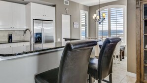 A modern kitchen with white cabinets, a stainless steel refrigerator, and a black countertop. There is a dining area with a chandelier and a view of the ocean through large windows.