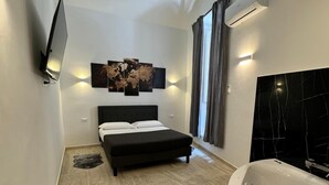 Une chambre moderne avec un grand lit, une tête de lit noire, un téléviseur fixé au mur et une grande fenêtre avec des rideaux.