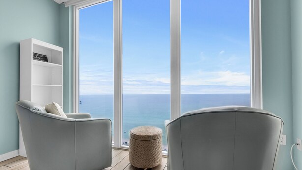 A room with a large window overlooking the sea, two grey armchairs, a white shelving unit, and a woven ottoman. - 12th fl Beachfront 3BR/3BA & Beach Service / Xtra Fridge & Icemaker Sleeps 8 (Navarre Beach)