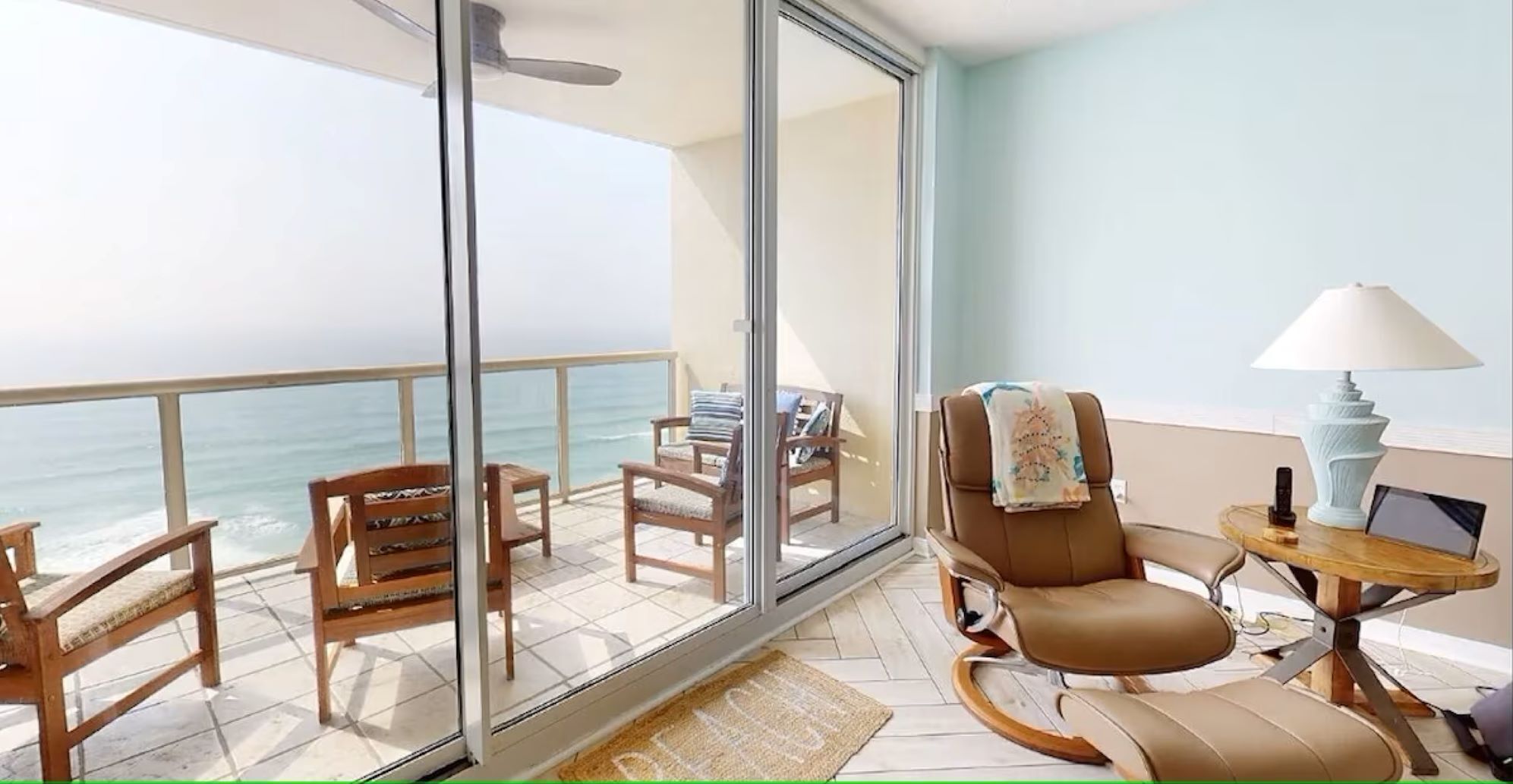 A balcony with a view of the ocean, furnished with wooden chairs and a leather sofa, and a small table with a lamp.