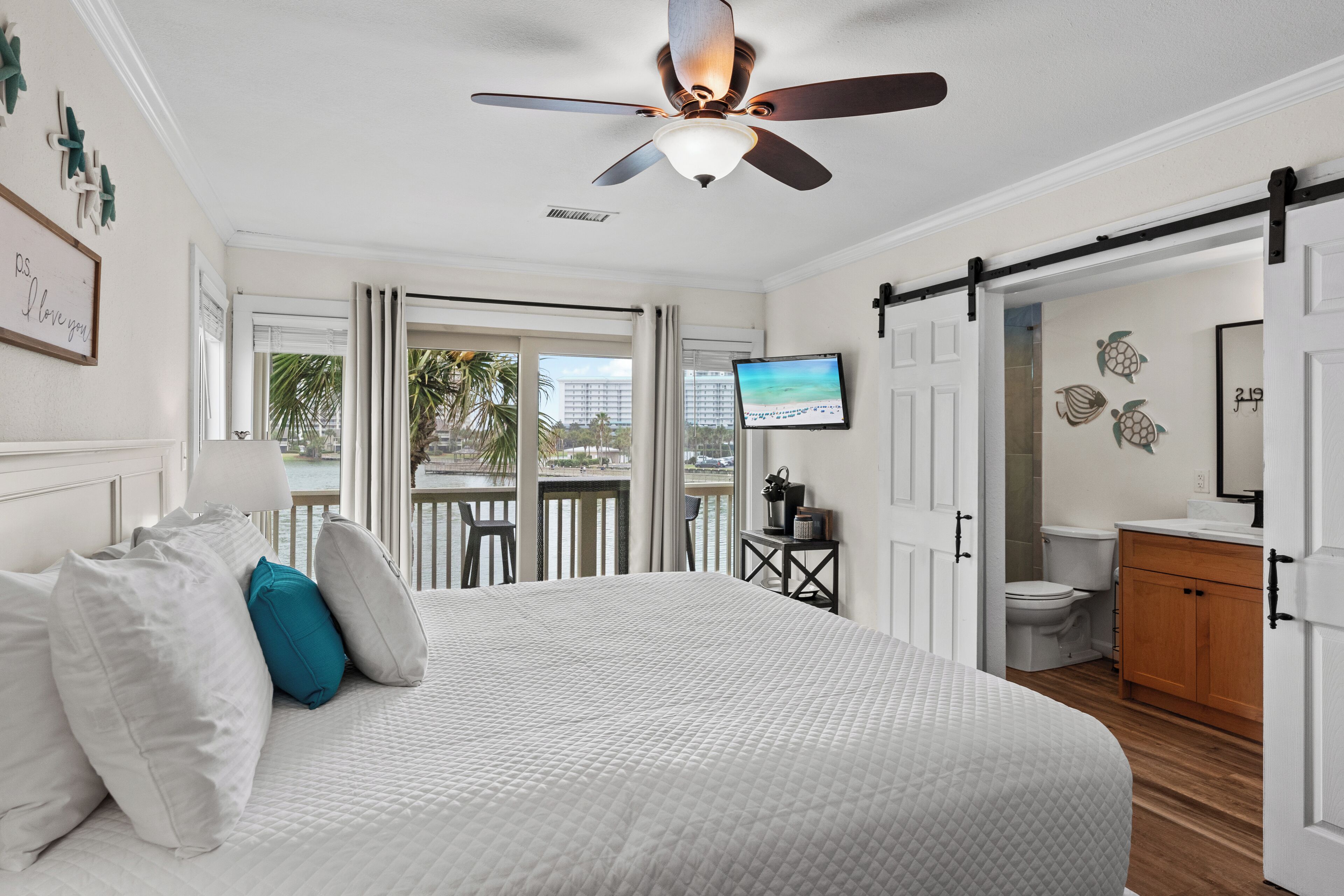 A hotel room with a bed, a ceiling fan, a TV, a balcony with a view of palm trees, and a bathroom with a toilet and sink.
