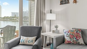 Ein Zimmer mit Blick auf ein Gewässer, ein grauer Sessel mit einem Kissen mit der Aufschrift 'BEACH', ein weißer Tisch mit einer Lampe und ein Wanddekor mit der Aufschrift 'welcome to PARADISE'.