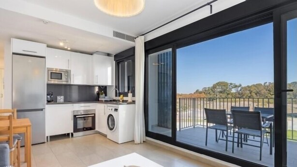 Eine moderne Küche mit weißen Schränken, einer Mikrowelle und einer Waschmaschine. Ein Balkon mit Sitzgelegenheit ist vorhanden.