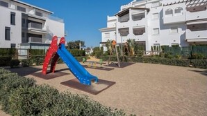 Ein Spielplatz mit einer blauen Rutsche und einer roten Kletterkonstruktion, umgeben von weißen Wohngebäuden.