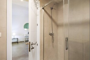 Un baño moderno con ducha de vidrio, una puerta blanca y un pasillo visible que conduce a una habitación con una mesa y una silla.