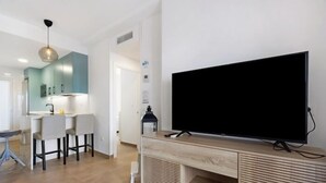 A modern living room with a large flat-screen TV mounted on a wooden console, a dining area with a white table and chairs, and a kitchen with teal cabinets and a hanging light fixture. - Isla Candor free parking by Staycare (Rota)