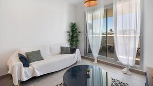 Een moderne woonkamer met een witte bank, een glazen salontafel en een groot raam met transparante gordijnen.