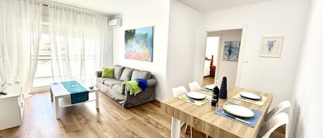 A modern living room with a dining area, a grey sofa with cushions, a wooden dining table set for four, and a painting on the wall.