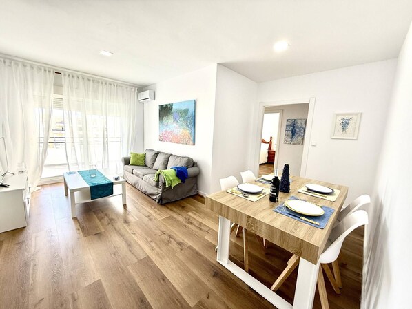 A modern living room with a dining area, a grey sofa with cushions, a wooden dining table set for four, and a painting on the wall.